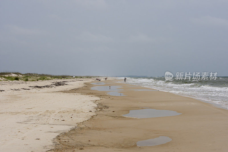 View along overcast beach with breaking surf and distant people fishing at low tide